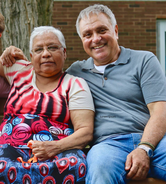 Picture10 - Toronto Bangla Town | Digital Bangla Weekly Magazine Two smiling adults seated outdoors, a woman in a colorful patterned dress and a man in a gray polo shirt, with the man's arm around her.