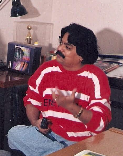 Picture11 - Toronto Bangla Town | Digital Bangla Weekly Magazine Man with mustache in a red-and-white striped sweater sits at a desk, gesturing with his hand while holding a remote.