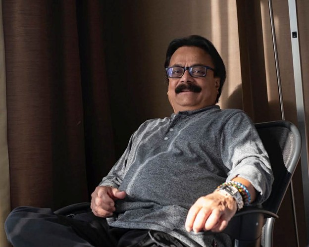 Picture11 - Toronto Bangla Town | Digital Bangla Weekly Magazine Man with a mustache and blue glasses sitting in an office chair, smiling at the camera in a relaxed pose.