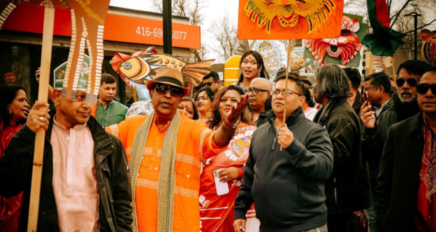 Picture12 - Toronto Bangla Town | Digital Bangla Weekly Magazine Participants in a colorful street parade, with one man in orange attire speaking into a microphone while others crowd around him.