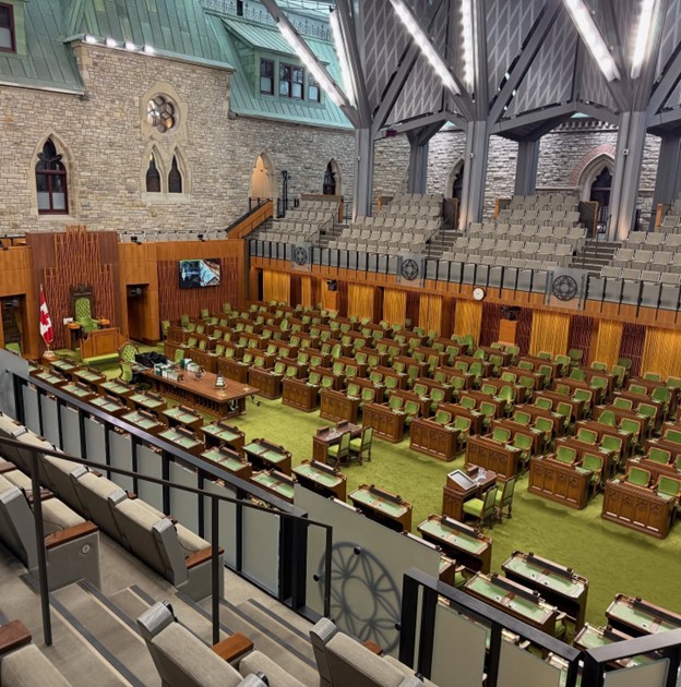 Picture3 - Toronto Bangla Town | Digital Bangla Weekly Magazine Legislative chamber with green upholstered seats, wooden desks, and a Canadian flag at the front; stone walls and a high vaulted ceiling.