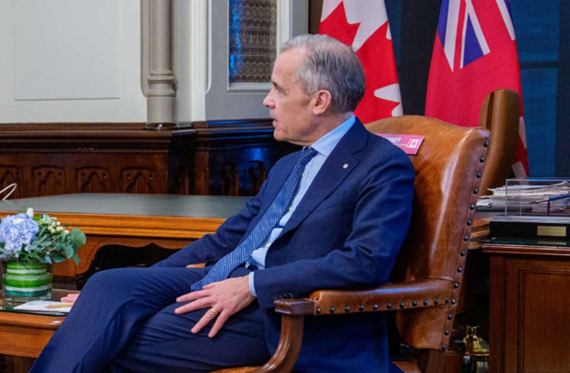 Picture3 - Toronto Bangla Town | Digital Bangla Weekly Magazine Side-profile of a man in a blue suit and tie seated in a brown leather chair in a formal office, with Canadian and other flags in the background.