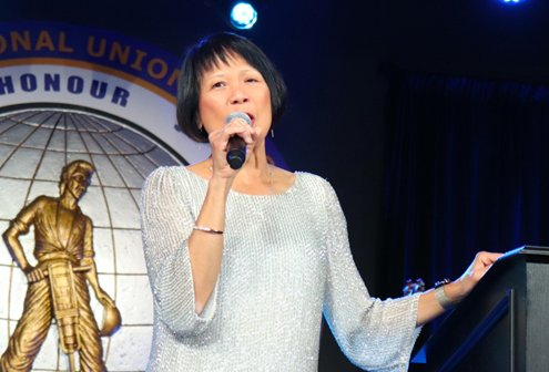 Picture6 - Toronto Bangla Town | Digital Bangla Weekly Magazine Woman in a silver dress speaks into a microphone on a lit stage, with a large seal and globe emblem in the background.