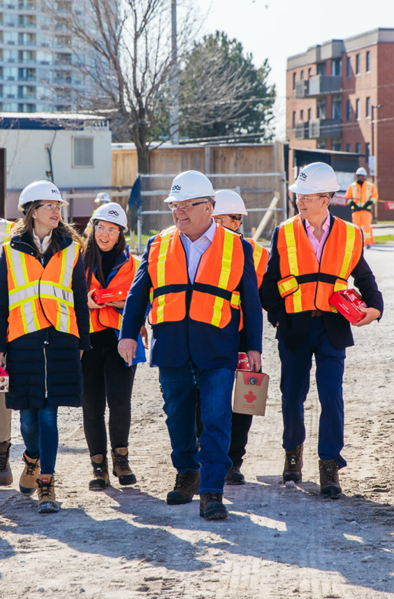 Picture6 - Toronto Bangla Town | Digital Bangla Weekly Magazine A group of construction professionals in hard hats and bright orange vests walking on a gravel site, discussing plans.
