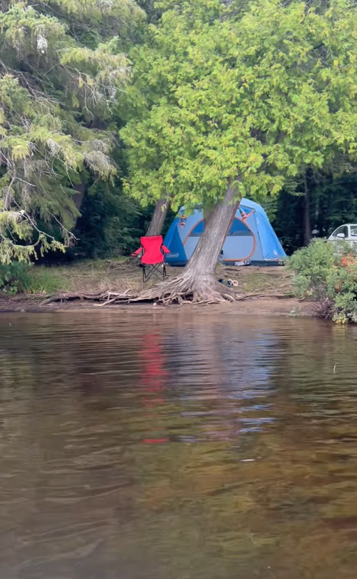 Picture7 - Toronto Bangla Town | Digital Bangla Weekly Magazine Blue tent set up on a lakeside shore behind a red camping chair among green trees, with water in the foreground and a car on the right edge.