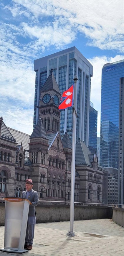 Picture9 - Toronto Bangla Town | Digital Bangla Weekly Magazine Man in traditional dress speaks at a podium on a rooftop; Nepali flag, historic clock-tower building, and modern skyscrapers in the background.