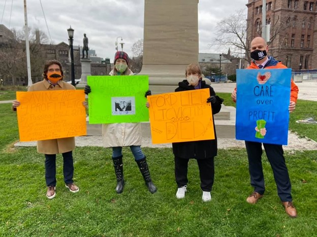 Picture1 - Toronto Bangla Town | Digital Bangla Weekly Magazine Four people wearing masks stand on a grassy area, each holding bright protest signs. From left to right: an orange sign; a neon green sign; an orange sign; and a blue sign reading 'CARE over PROFIT' with a small drawing. A statue and brick buildings are in the background.