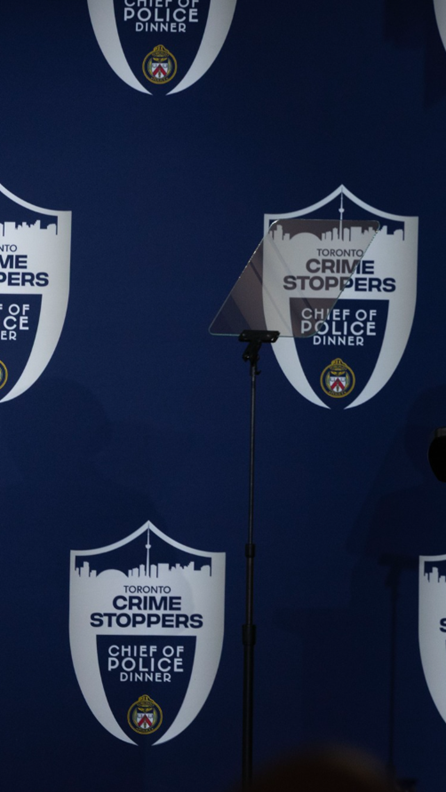 Picture1 - Toronto Bangla Town | Digital Bangla Weekly Magazine Stage backdrop for a Toronto Crime Stoppers Chief of Police Dinner, featuring shield logos and a microphone on a stand center stage.