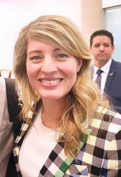 Picture10 - Toronto Bangla Town | Digital Bangla Weekly Magazine Smiling blonde woman in a plaid blazer in the foreground, with a suited man blurred in the background.