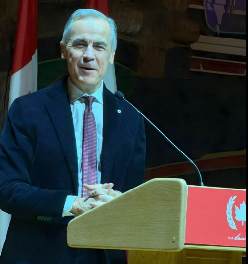 Picture10 - Toronto Bangla Town | Digital Bangla Weekly Magazine Man in a dark suit and red tie speaks at a wooden podium, with a Canadian flag visible in the background.