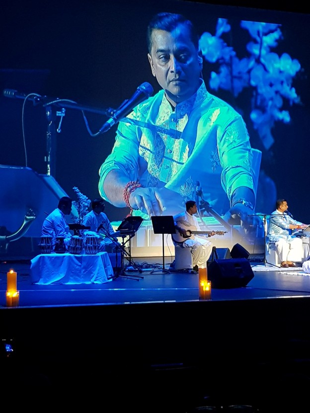 Picture10 - Toronto Bangla Town | Digital Bangla Weekly Magazine Live stage performance with musicians in white, guitarist center, large screen projection of a man's portrait behind; candles on stage.