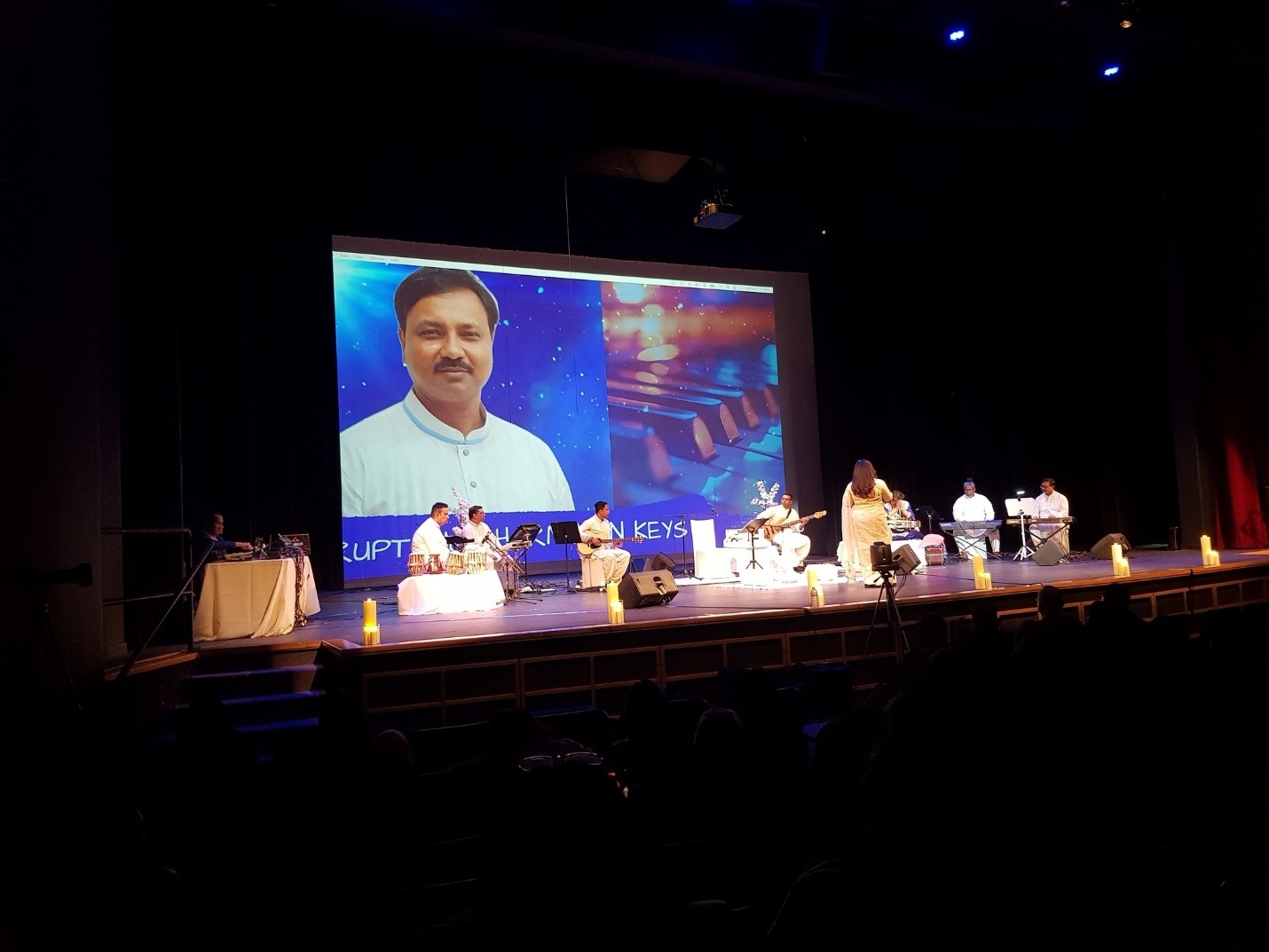 Picture11 - Toronto Bangla Town | Digital Bangla Weekly Magazine Stage performance with a large screen showing a man's portrait; musicians sit with instruments as a conductor leads.