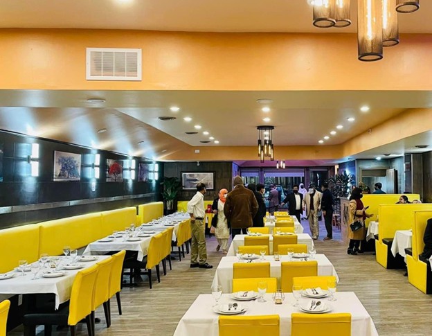 Picture11 - Toronto Bangla Town | Digital Bangla Weekly Magazine Restaurant dining room with yellow chairs and white-tablecloth tables; guests stand and chat in a warmly lit hall.