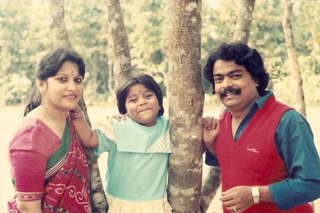 Picture11 - Toronto Bangla Town | Digital Bangla Weekly Magazine Family of three posing between tree trunks in a park; mother in a pink saree, daughter in turquoise dress, father in red sweater smiling at the camera.