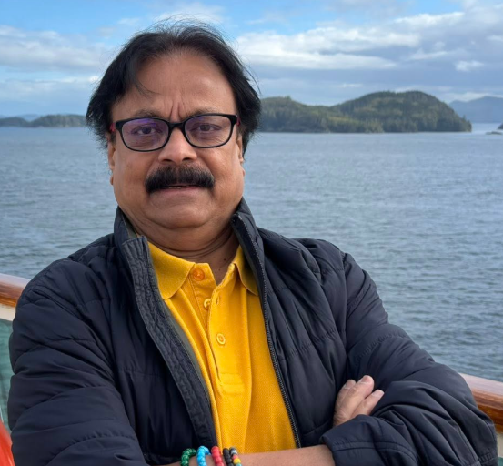 Picture11 - Toronto Bangla Town | Digital Bangla Weekly Magazine Man with glasses and mustache, wearing a yellow polo and dark jacket, stands with arms crossed on a boat railing with water and distant islands behind him.