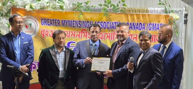 Picture12 - Toronto Bangla Town | Digital Bangla Weekly Magazine Six men in suits pose on a stage as one hands a certificate to another, with a microphone in hand and a GMAC banner in the background.