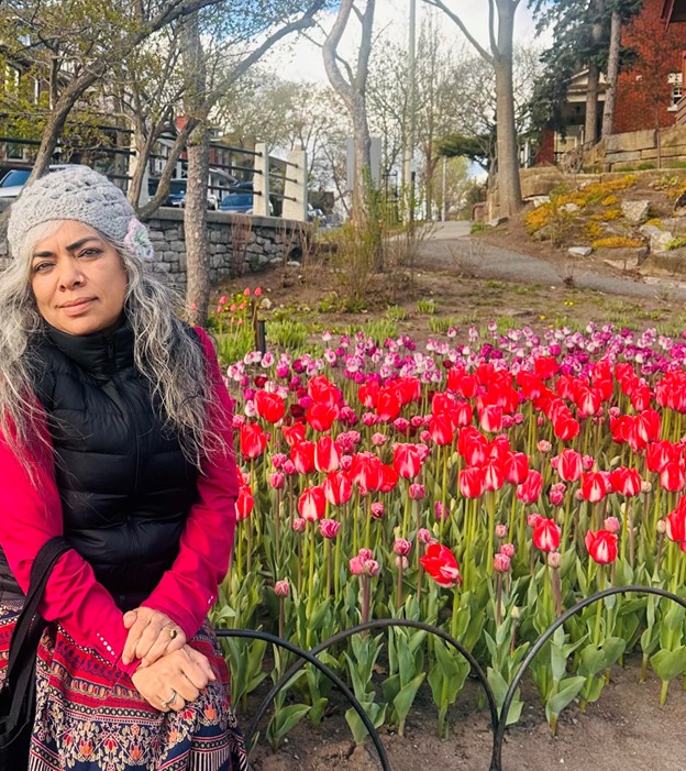 Picture12 - Toronto Bangla Town | Digital Bangla Weekly Magazine Woman with gray hair and a knit hat sits beside a bed of red and pink tulips in a park, wearing a pink jacket and black vest.