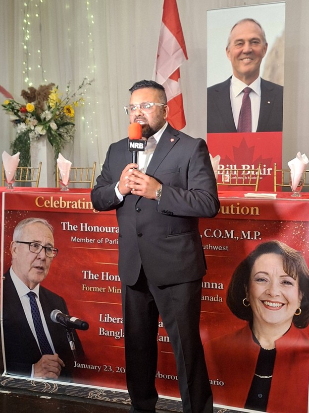 Picture12 - Toronto Bangla Town | Digital Bangla Weekly Magazine Man in a dark suit and glasses speaks into an orange NRB microphone at a formal event, standing before a red banner with portraits of politicians.