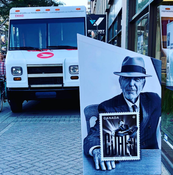 Picture2 - Toronto Bangla Town | Digital Bangla Weekly Magazine Canada Post delivery van behind a large cutout of a suited man wearing a fedora, holding a stamp image on a city sidewalk.