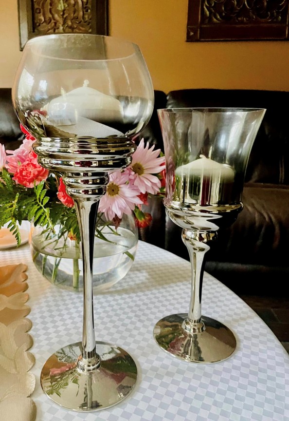 Picture3 - Toronto Bangla Town | Digital Bangla Weekly Magazine Two tall silver candle holders with glass bowls sit on a checkered tablecloth, a vase of pink flowers behind them—a decorative centerpiece.