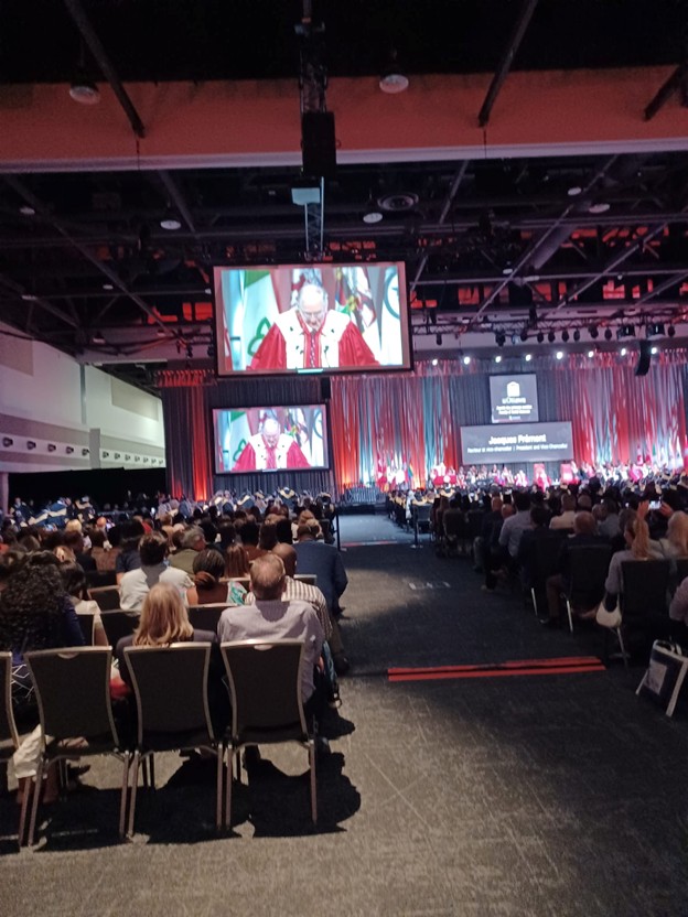 Picture3 - Toronto Bangla Town | Digital Bangla Weekly Magazine Large conference hall with an audience facing a decorated stage; two screens show a speaker in a red robe.