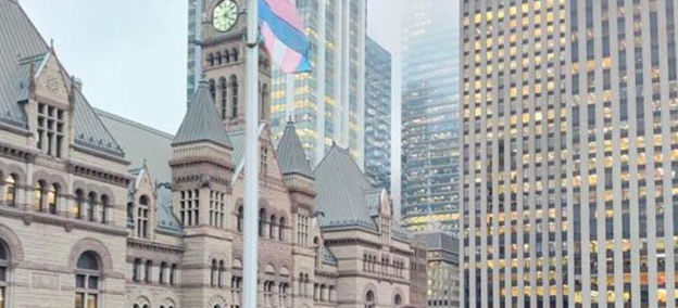 Picture4 - Toronto Bangla Town | Digital Bangla Weekly Magazine Historic stone building with turrets and a clock tower in front of modern glass skyscrapers in a downtown skyline, with a flagpole nearby.
