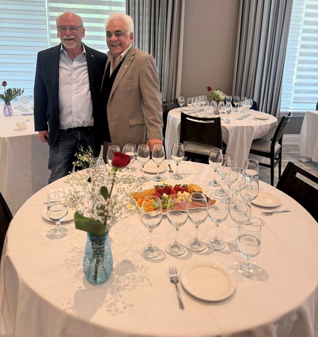 Picture5 - Toronto Bangla Town | Digital Bangla Weekly Magazine Two smiling older men in jackets pose beside a round banquet table set with multiple wine glasses and a floral centerpiece in a formal dining room