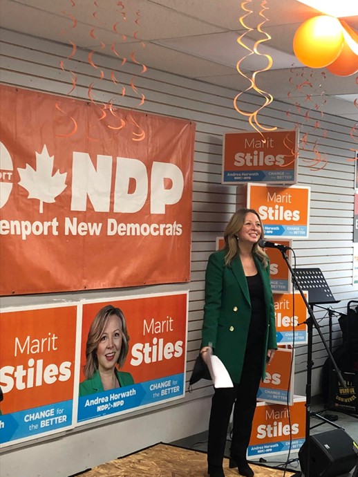 Picture7 - Toronto Bangla Town | Digital Bangla Weekly Magazine Woman in a green blazer stands at a microphone during a campaign event, with orange NDP banners and posters that read 'Marit Stiles' and 'Change for the Better' in the background.