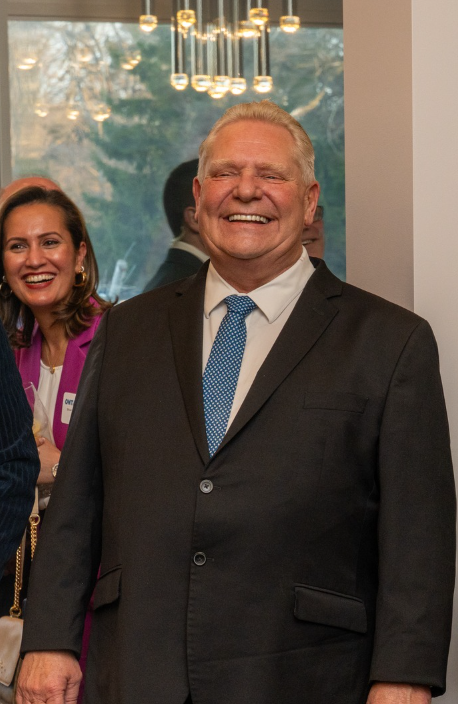 Picture9 - Toronto Bangla Town | Digital Bangla Weekly Magazine Smiling older man in a dark suit and blue tie at an indoor event, with a woman in a purple jacket behind him.