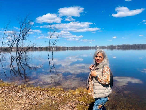 saji - Toronto Bangla Town | Digital Bangla Weekly Magazine Woman with gray hair standing by a calm lake, trees reflecting in the water, blue sky and clouds above, wearing a puffer vest and patterned blouse.
