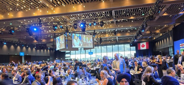 Picture1 - Toronto Bangla Town | Digital Bangla Weekly Magazine Large conference hall crowded with attendees at round tables; screens show speakers and a Canadian flag is visible in the background.