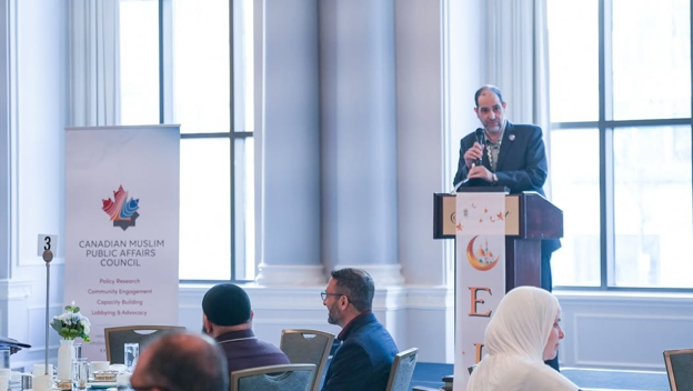 Picture1 - Toronto Bangla Town | Digital Bangla Weekly Magazine Man in a suit speaks at a podium during a conference; audience sits at tables, banner on the left shows the Canadian Muslim Public Affairs Council logo and name.