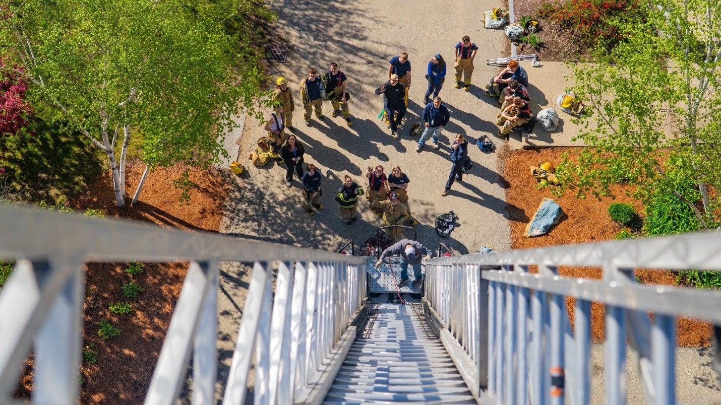Picture10 - Toronto Bangla Town | Digital Bangla Weekly Magazine Aerial view of firefighters and trainees looking up at a firefighter descending a tall ladder from a tower.
