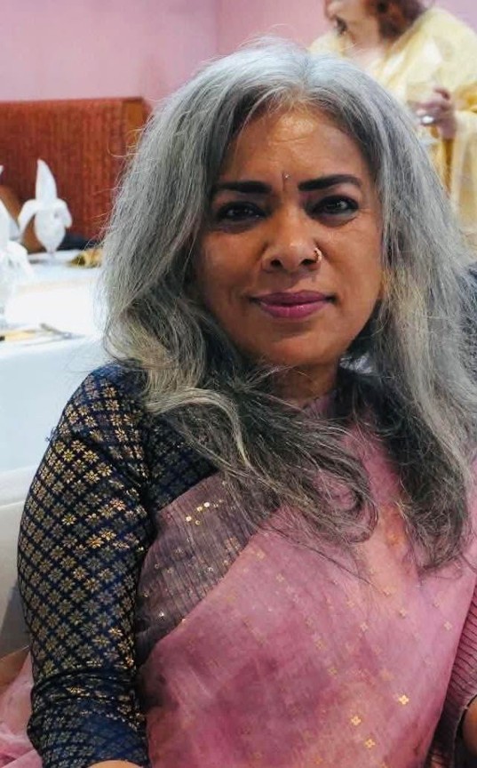 Picture11 - Toronto Bangla Town | Digital Bangla Weekly Magazine Smiling woman with long gray hair wearing a pink saree with black patterned sleeves, seated at a banquet table in a hall.