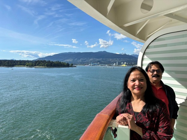 Picture11 - Toronto Bangla Town | Digital Bangla Weekly Magazine Two smiling adults lean on a cruise ship balcony railing, overlooking calm blue water with an island and distant mountains in the background.