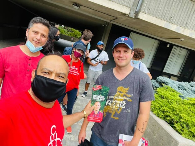 Picture11 - Toronto Bangla Town | Digital Bangla Weekly Magazine Group of volunteers outside a modern building, with a man in a red shirt wearing a black mask handing a flyer to a man in a gray shirt and blue cap who is receiving it; several people in the background wearing masks.
