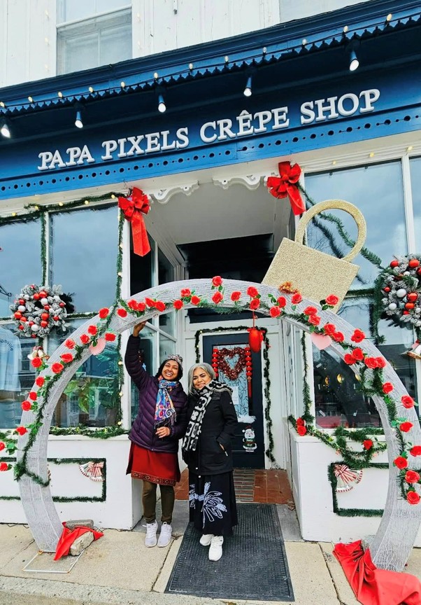 Picture12 - Toronto Bangla Town | Digital Bangla Weekly Magazine Two women pose inside a white arch adorned with red roses in front of a blue storefront reading 'PAPA PIXELS CRÊPE SHOP', with festive holiday decorations.