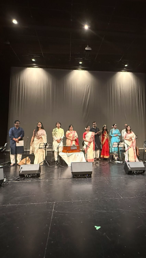 Picture12 - Toronto Bangla Town | Digital Bangla Weekly Magazine Group of performers in traditional Indian attire standing on a stage with music stands and instruments under bright spotlights, curtain backdrop behind them.