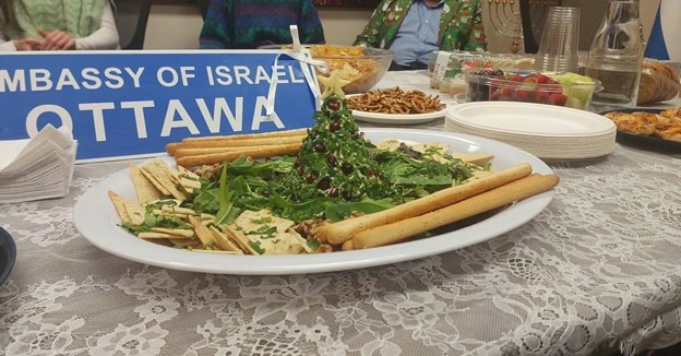Picture2 - Toronto Bangla Town | Digital Bangla Weekly Magazine Plate of assorted crackers and greens with a central veggie garnish on a lace-covered table, a blue 'Embassy of Israel Ottawa' sign visible in the background.