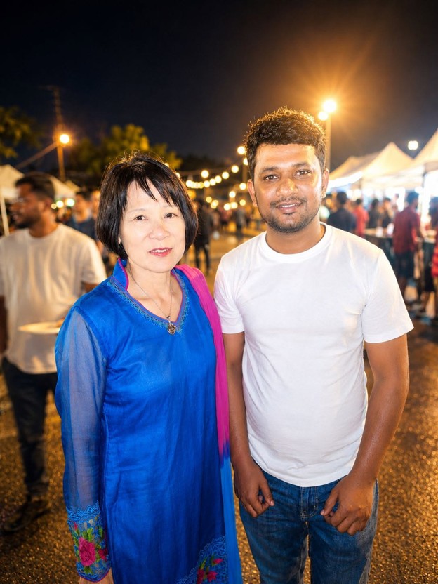 Picture2 - Toronto Bangla Town | Digital Bangla Weekly Magazine Two people posing together at a nighttime market with string lights and vendor stalls in the background; a woman in a blue traditional outfit stands beside a man in a white t‑shirt.