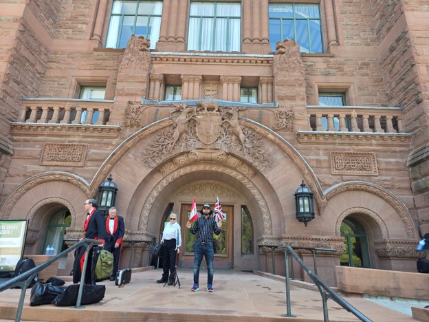 Picture2 - Toronto Bangla Town | Digital Bangla Weekly Magazine Group of people standing on the steps of a large ornate red-brick building with circular archway and carved coats of arms; a man in a striped shirt holds up two flags while others watch, luggage at their feet.