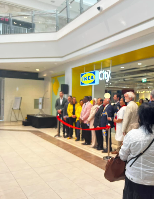 Picture2 - Toronto Bangla Town | Digital Bangla Weekly Magazine Group of people in business and casual attire standing behind a red ribbon at an IKEA City store opening in a mall atrium.