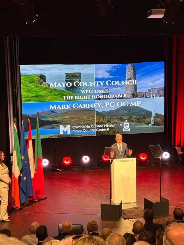 Picture3 - Toronto Bangla Town | Digital Bangla Weekly Magazine Speaker at podium on stage with a large screen behind him announcing Mark Carney's welcome by Mayo County Council; flags line the left side, audience in foreground.