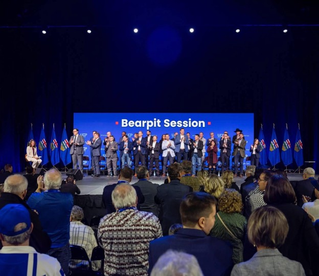 Picture3 - Toronto Bangla Town | Digital Bangla Weekly Magazine Group of people standing on a stage during a Bearpit Session event, with a blue backdrop and flags on both sides, audience watching from the hall.