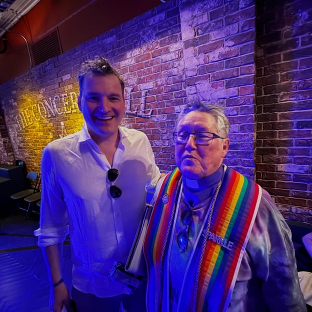 Picture3 - Toronto Bangla Town | Digital Bangla Weekly Magazine Two men pose for a photo in a brick-walled venue with blue lighting; the man on the left wears a white shirt with sunglasses hanging from his chest, while the man on the right wears a rainbow Pride sash and glasses.