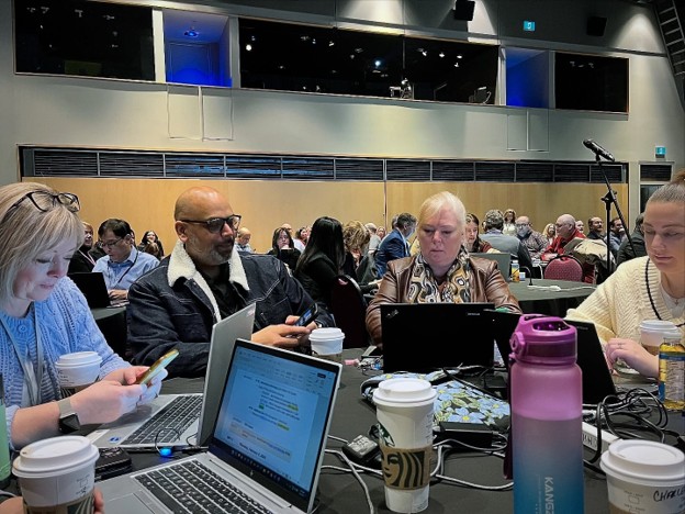 Picture3 - Toronto Bangla Town | Digital Bangla Weekly Magazine Group of professionals seated around a conference table with laptops, coffee cups, and a water bottle in a large meeting hall of attendees.