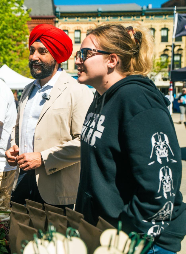 Picture3 - Toronto Bangla Town | Digital Bangla Weekly Magazine Man wearing a red turban and beige blazer stands beside a woman in a Star Wars hoodie at an outdoor market/wairing stalls in the foreground.