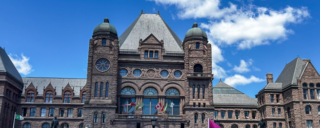 Picture4 - Toronto Bangla Town | Digital Bangla Weekly Magazine Front view of a historic red-brick building with two rounded towers, arched windows, and flags on the facade against a blue sky.