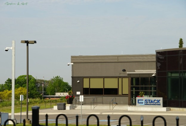 Picture6 - Toronto Bangla Town | Digital Bangla Weekly Magazine Exterior view of a modern brown commercial building with a glass entrance and a blue-and-white Stack sign, surrounded by a paved parking area, lamp posts, and some greenery against a blue sky.