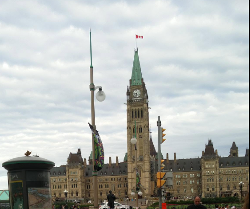 Picture7 - Toronto Bangla Town | Digital Bangla Weekly Magazine Ottawa's Parliament Hill with the Peace Tower, a Canadian flag, and historic stone buildings under a cloudy sky.